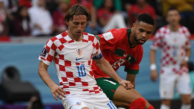 Modric e jogador marroquinho brigam pela bola durante partida. Eles vestem camisa branca com quadrados vermelhos e vermelha com short verde. Croacia e Marrocos disputam 3º lugar na copa